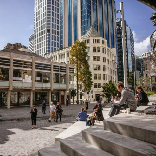 People sitting in the sun on the terracing in Freyberg Place in Auckland's city centre, with the statue of General Freyberg and the Ellen Melville Centre visible. Image: Sacha Stejko