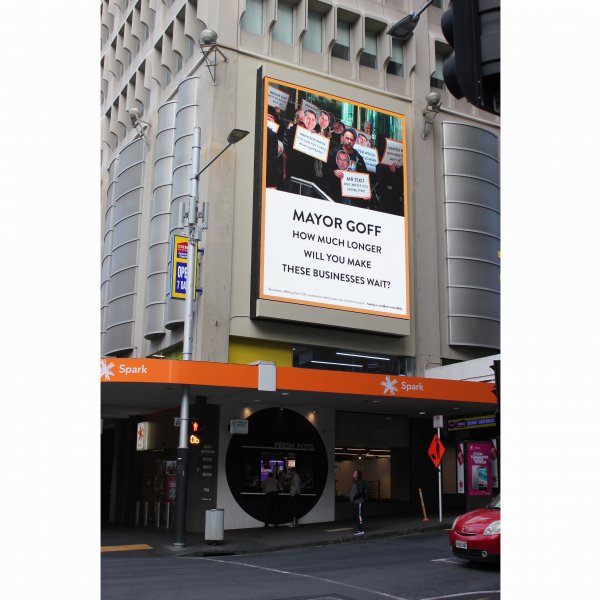 A large billboard on a building at the corner of Queen and Wyndham Streets, with the text 'Mayor Goff: how much longer will you make these businesses wait?'. Above the text is a photograph of business owners gathering in protest with signs.