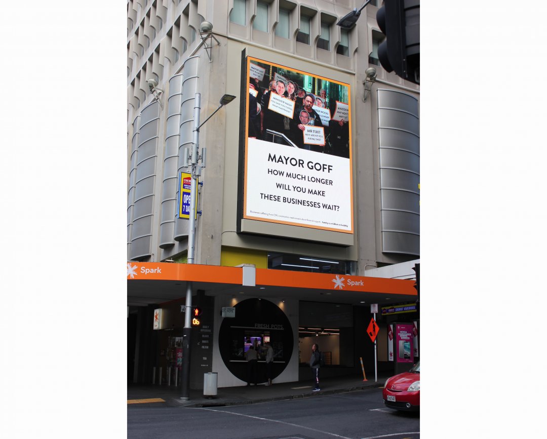 A large billboard on a building at the corner of Queen and Wyndham Streets, with the text 'Mayor Goff: how much longer will you make these businesses wait?'. Above the text is a photograph of business owners gathering in protest with signs.